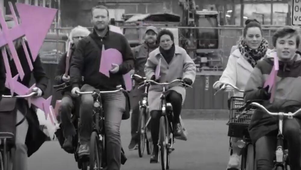 Verschillende mensen fietsen richting de camera. Ze houden elk een roze houten pijl vast.