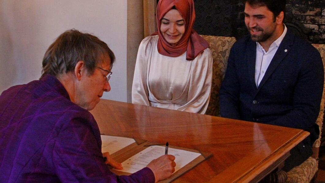 Marlies zet een handtekening op de trouwakte van Huseyin en Betül, die lachend toekijken