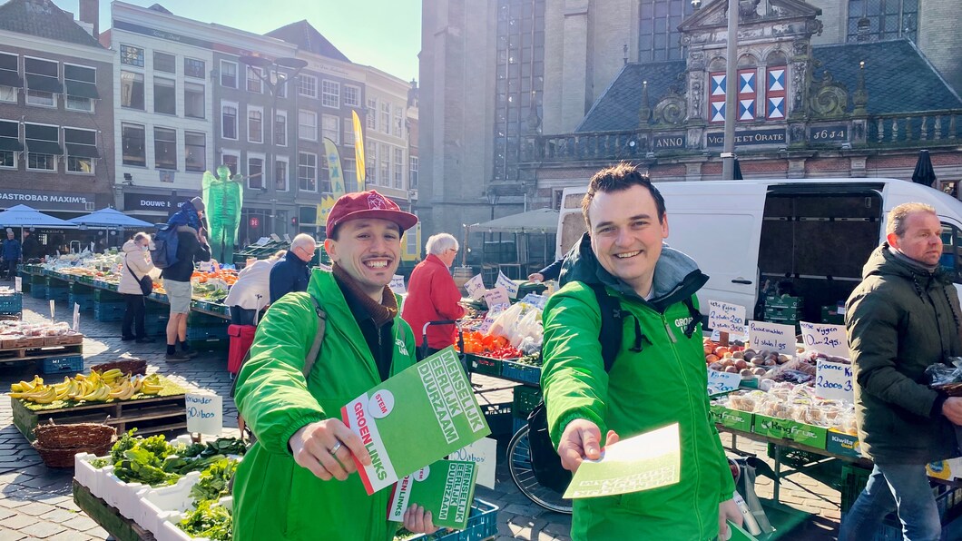 Joey en Neal hebben beide een GroenLinks-jasje aan. Ze staan in de stad, en steken allebei een flyer uit richting de camera.