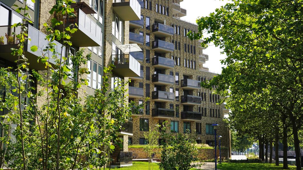 Een flat met balkons. Voor de flat staan bomen en planten rond een groen gazon.