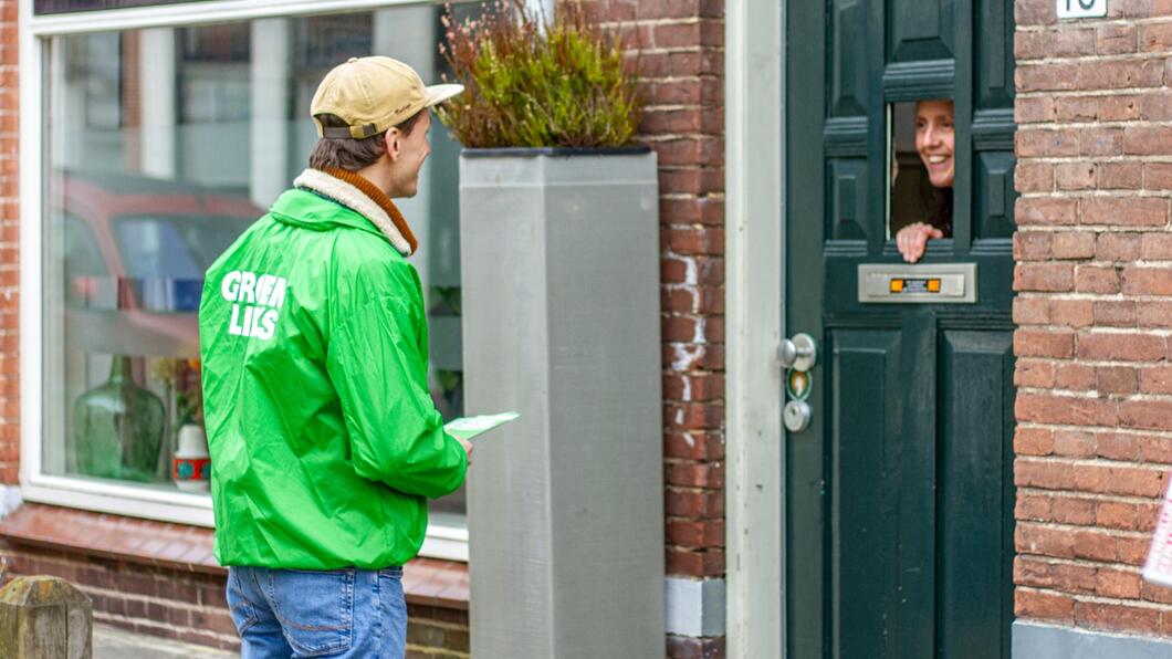 Een GroenLinks-vrijwilliger met een groen jasje aan staat voor een huis. De bewoner kijkt via een klein luikje in de voordeur naar buiten en lacht
