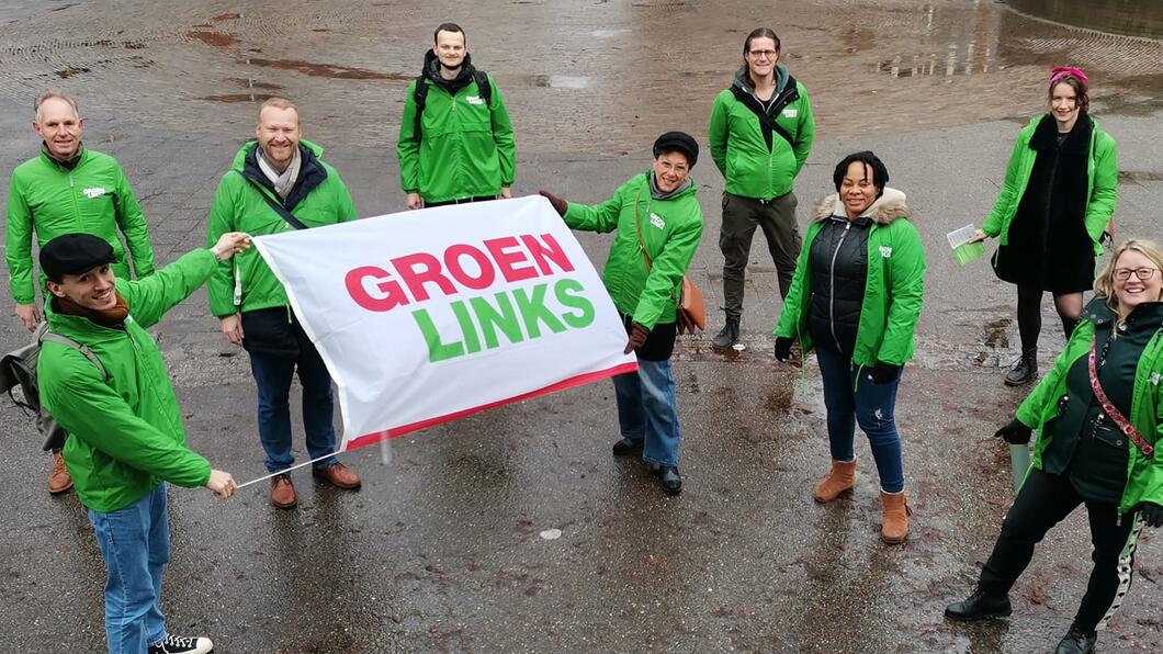 Een groep vrijwilligers met groene jasjes aan. Twee van hen houden een GroenLinks-vlag omhoog