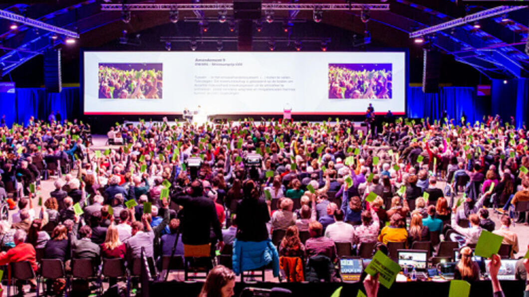 Een GroenLinks-congres, foto genomen vanuit achterin de zaal. Er zitten veel mensen in de zaal, vooraan hangen grote schermen.