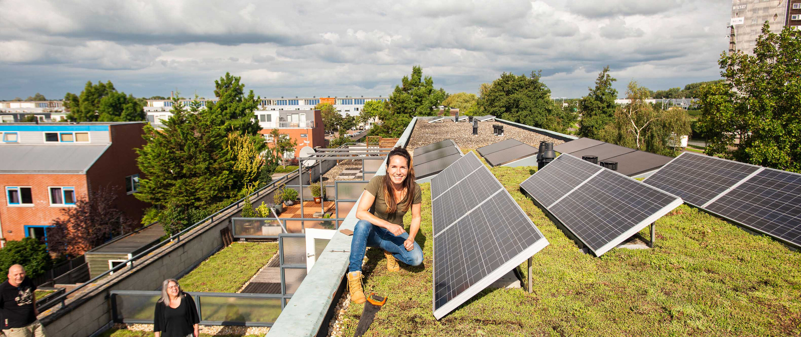 Een woonwijk met groene daken en zonnepanelen. Een vrouw zit naast een zonnepaneel en lacht naar de camera.