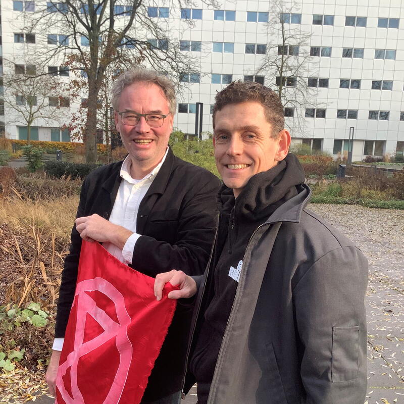 Martin en Rob houden een rode vlag met het symbool van XR erop vast. Ze zijn van de zijkant gefotografeerd.