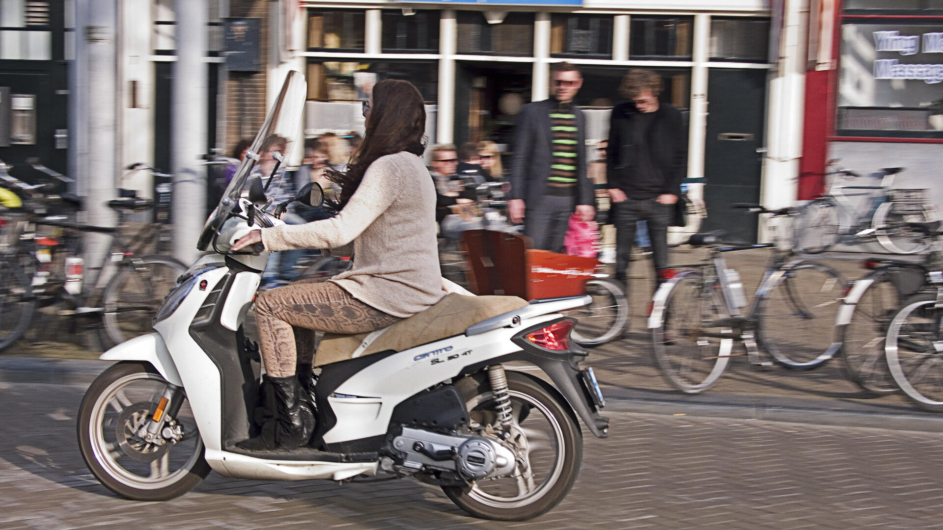 Een jonge vrouw rijdt op een scooter door een straat. Op de achtergrond staan twee mannen op de stoep, met om zich heen geparkeerde fietsen.