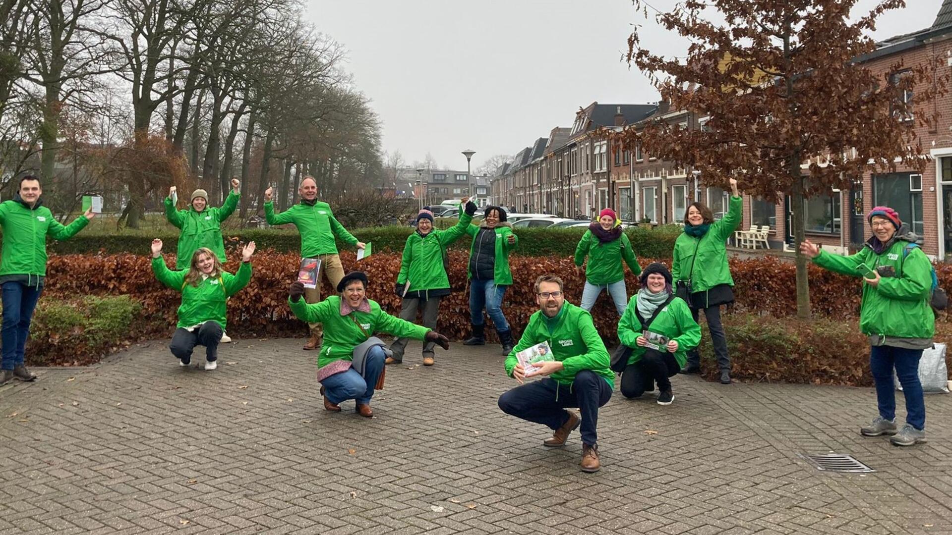 Een groep vrijwilligers met groene jasjes aan. Ze lachen en houden hun armen in de lucht