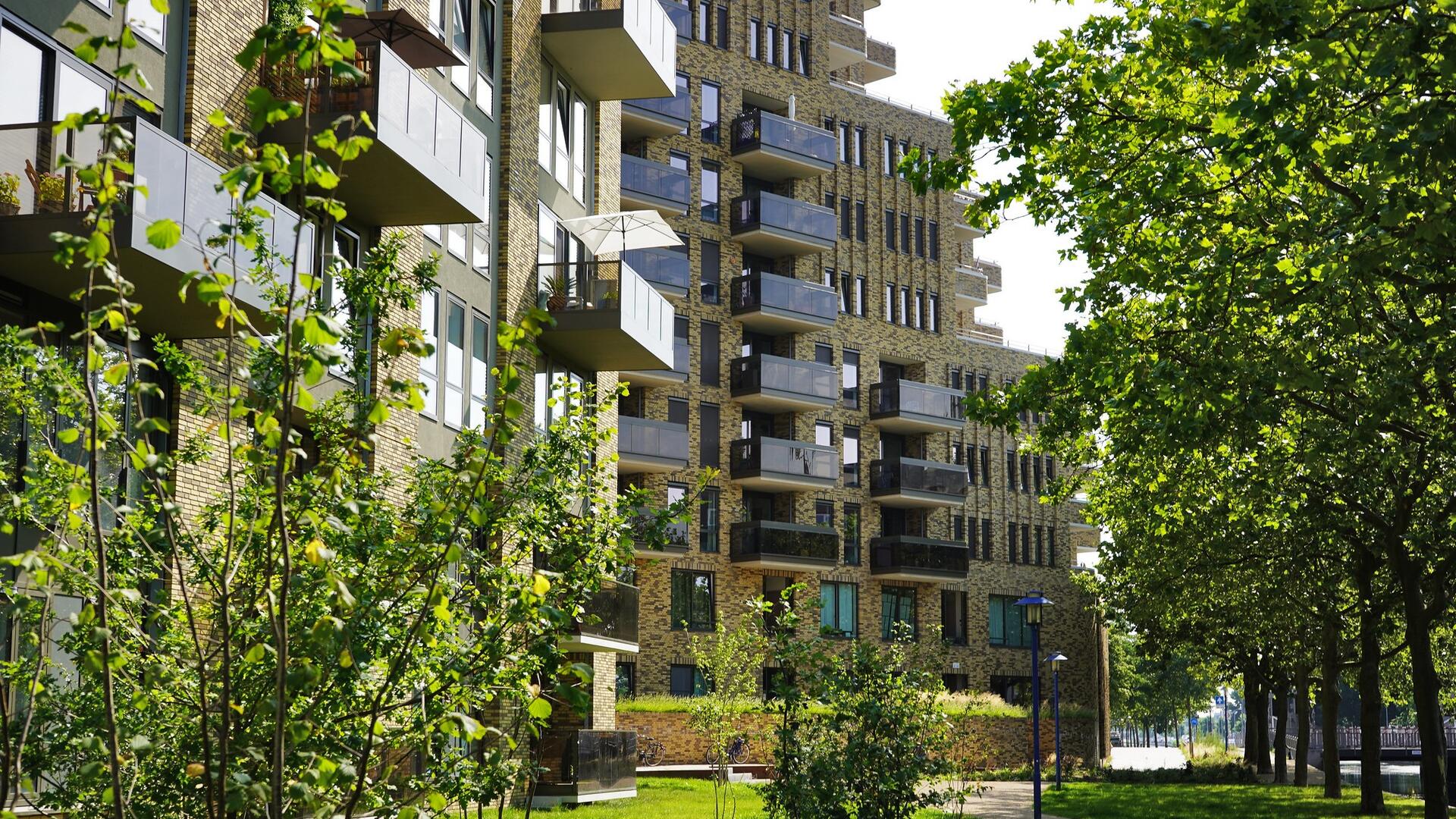 Een flat met balkons. Voor de flat staan bomen en planten rond een groen gazon.