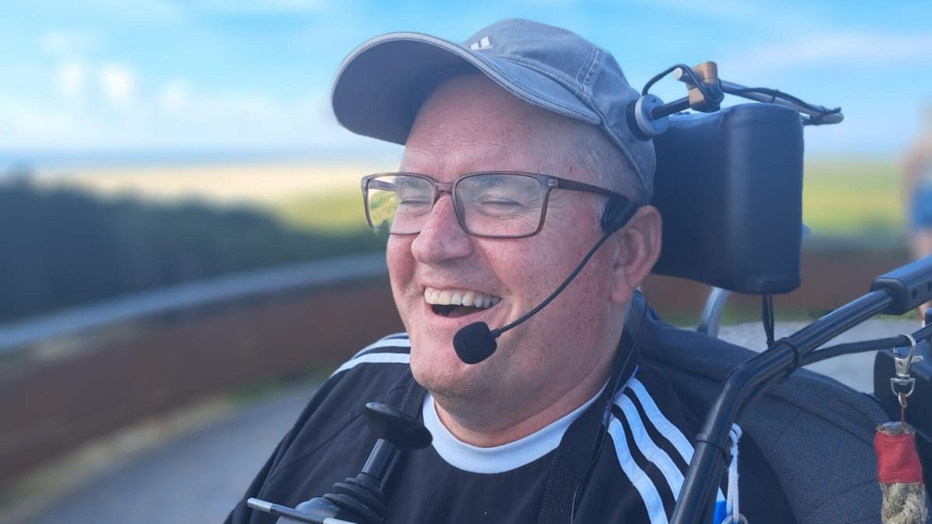 Geert Jan den Hengst lacht. Hij heeft een petje en een bril op en zit in een rolstoel.