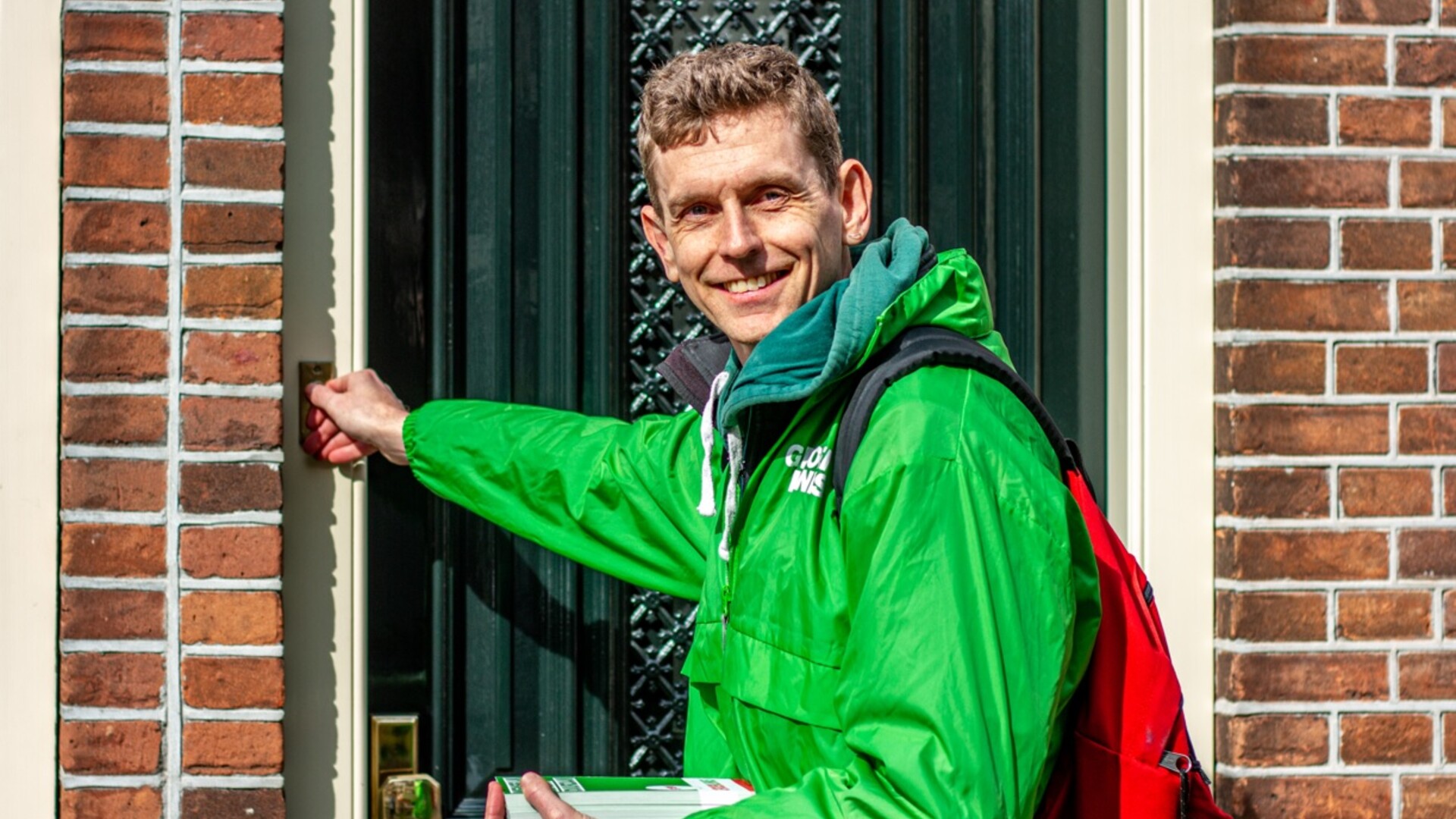 Een vrijwilliger met een groen jasje aan belt aan bij een voordeur.